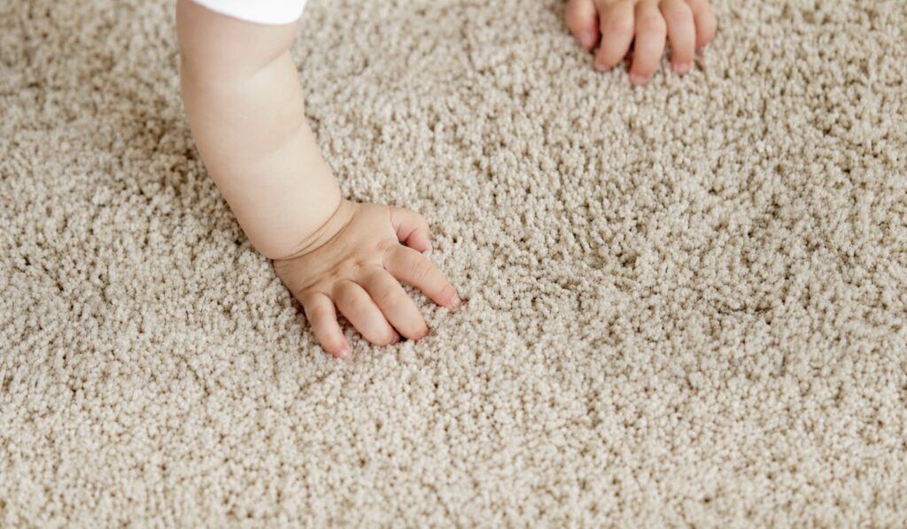 Baby crawling on a beige carpet with hands pressed into the fibers, illustrating how children can come into contact with household dust.