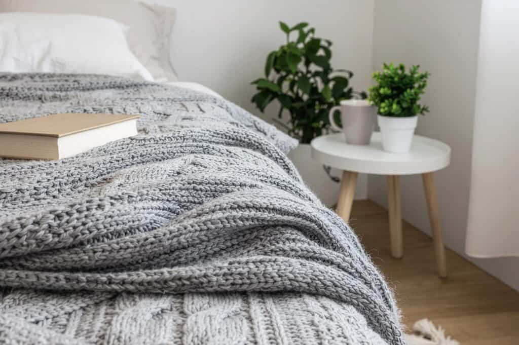 Cozy bedroom with a bed covered in a gray knit blanket, a book on the bed, and a small white side table with a mug and potted plants