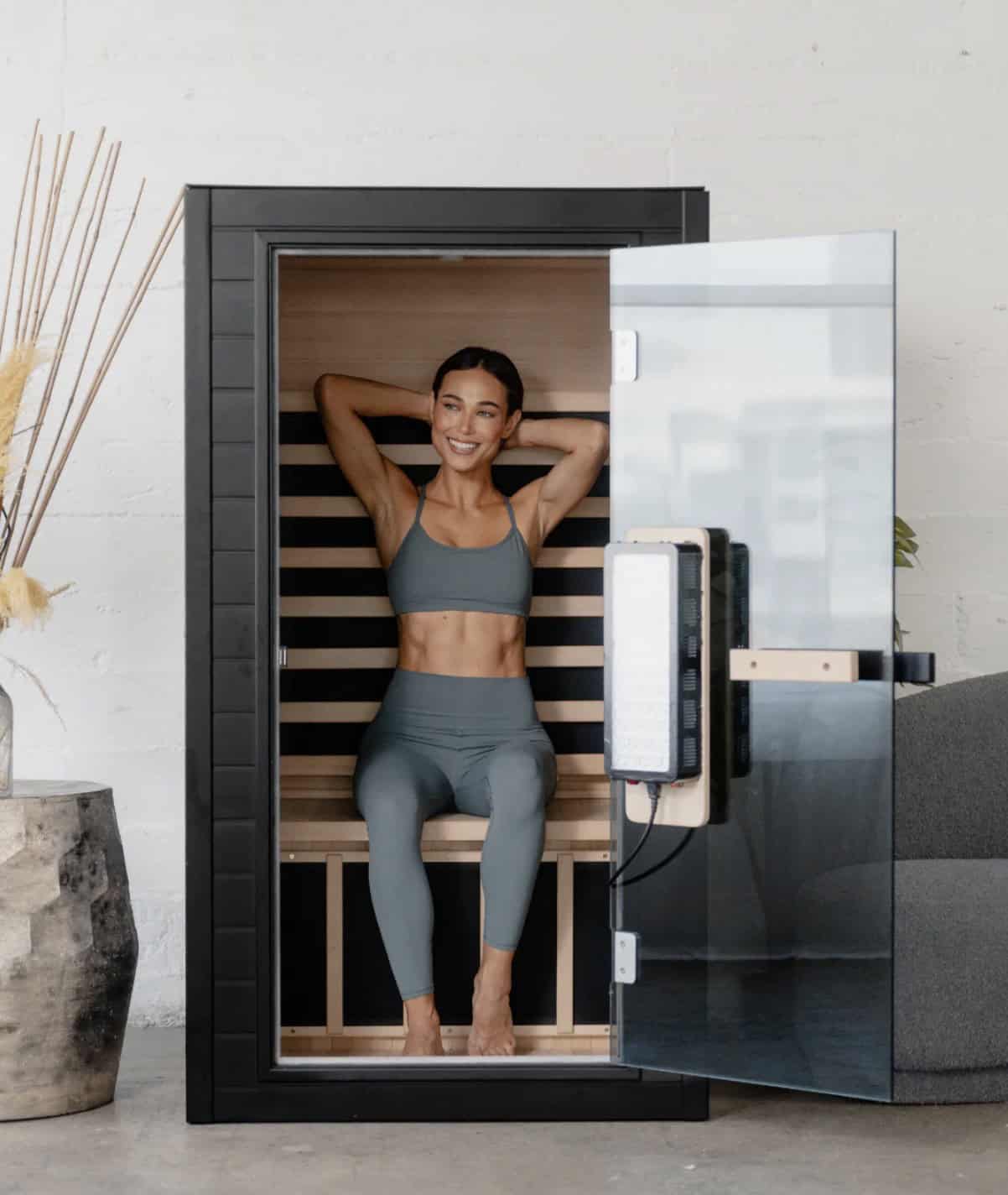 Person sitting inside a compact indoor infrared sauna with wooden interior and open glass door