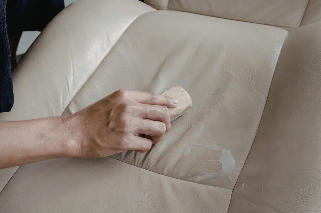 Hand scrubbing a light-colored faux leather couch cushion with visible wear and peeling surface.