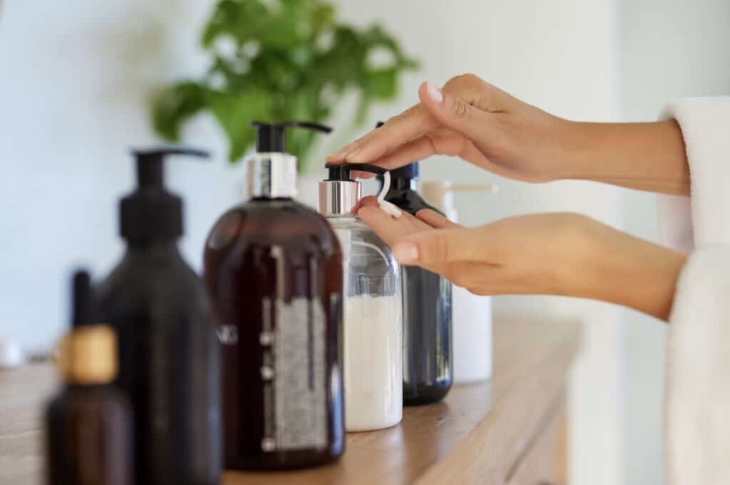 Close-up of hands dispensing lotion from a pump bottle on a wooden bathroom counter, with several reusable glass and plastic containers and a blurred plant in the background.