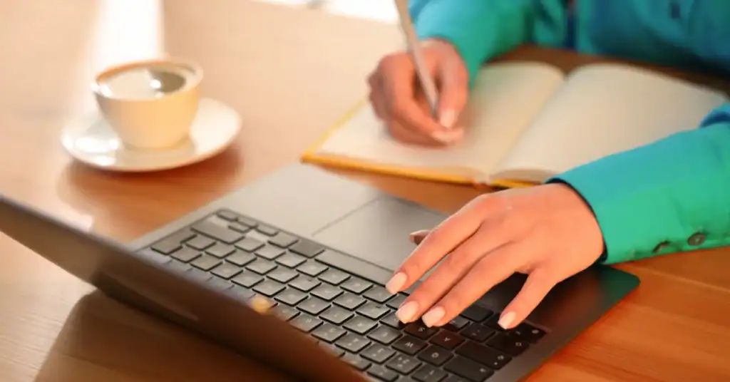 Hands using a laptop while writing notes in a notebook beside a coffee cup.