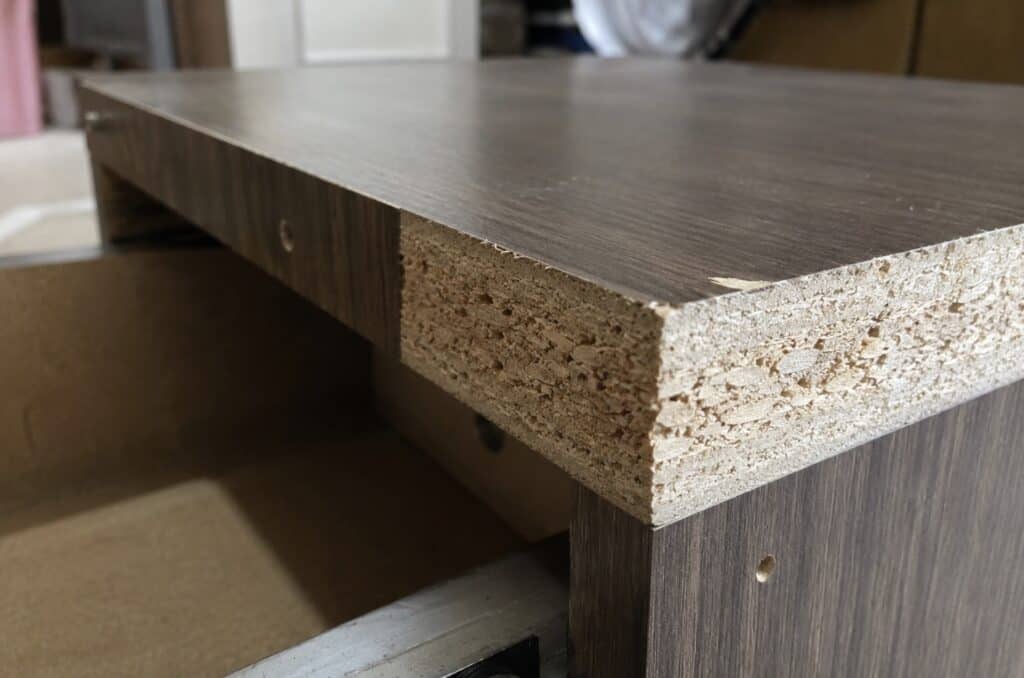 Close-up of damaged particleboard furniture showing an exposed engineered wood core beneath a white laminate surface.