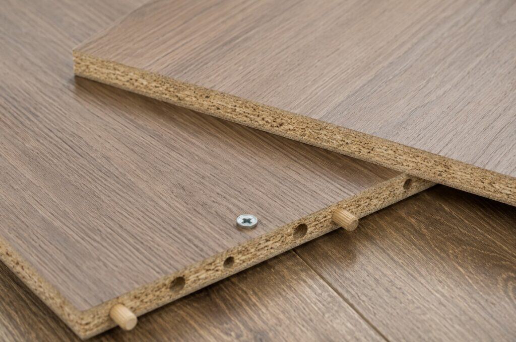 Close-up of particle board furniture panels with wood-look laminate surfaces, showing exposed compressed wood edges, pre-drilled holes, and assembly dowels in a realistic indoor setting.
