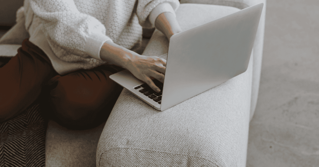 Person uses a laptop while sitting on a light fabric sofa to compare furniture options online.