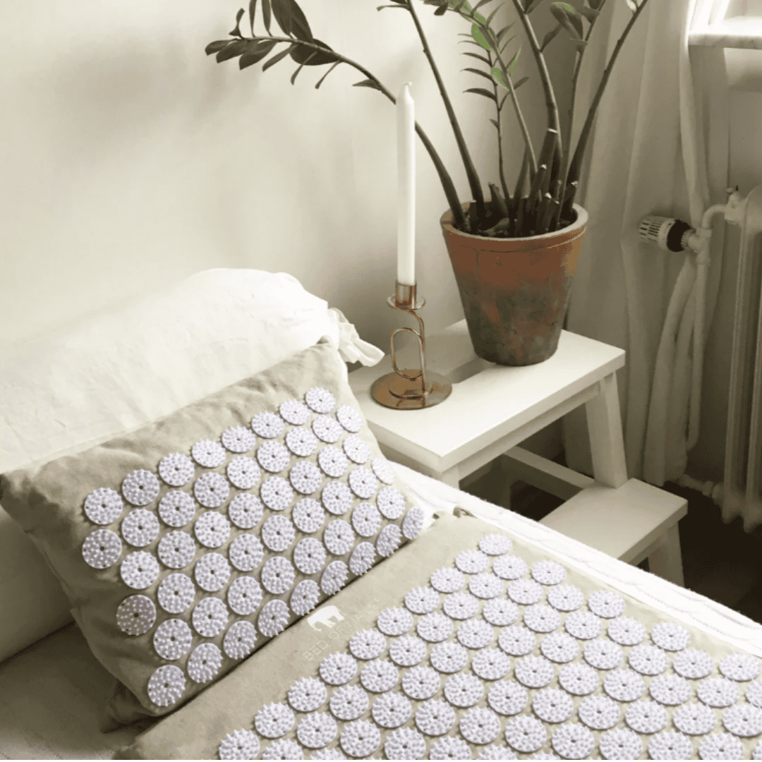 White acupressure mat and matching pillow arranged on a bed beside a plant and bedside table.