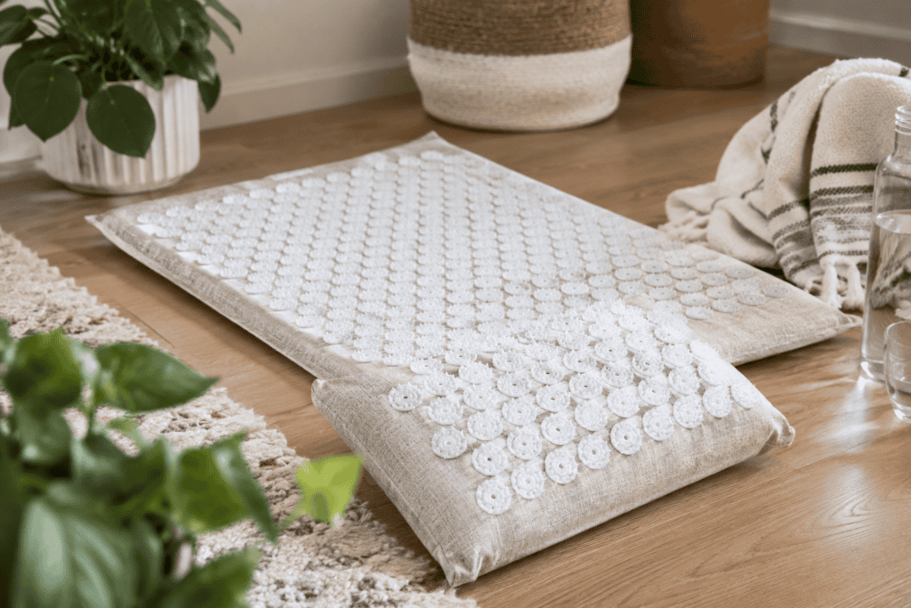 Natural linen acupressure mat and matching pillow with white pressure-point discs arranged on a wooden floor in a cozy, softly lit home setting.