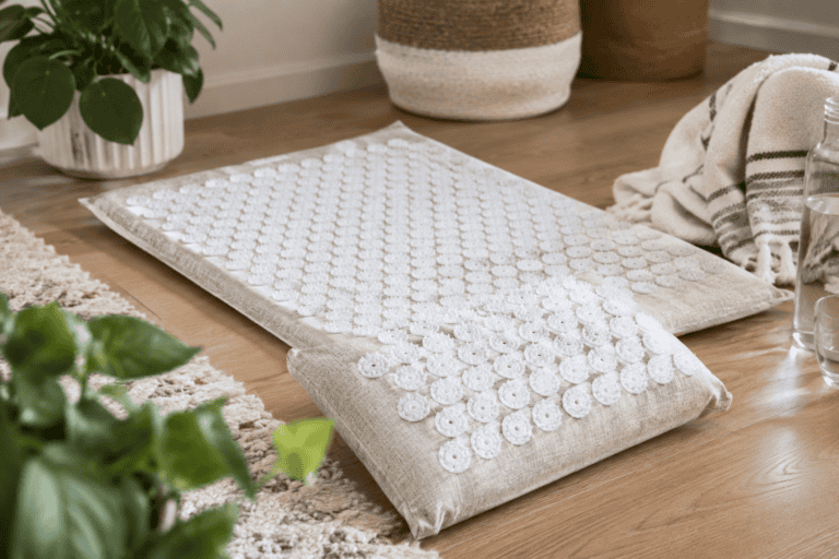 Natural linen acupressure mat and matching pillow with white pressure-point discs arranged on a wooden floor in a cozy, softly lit home setting.
