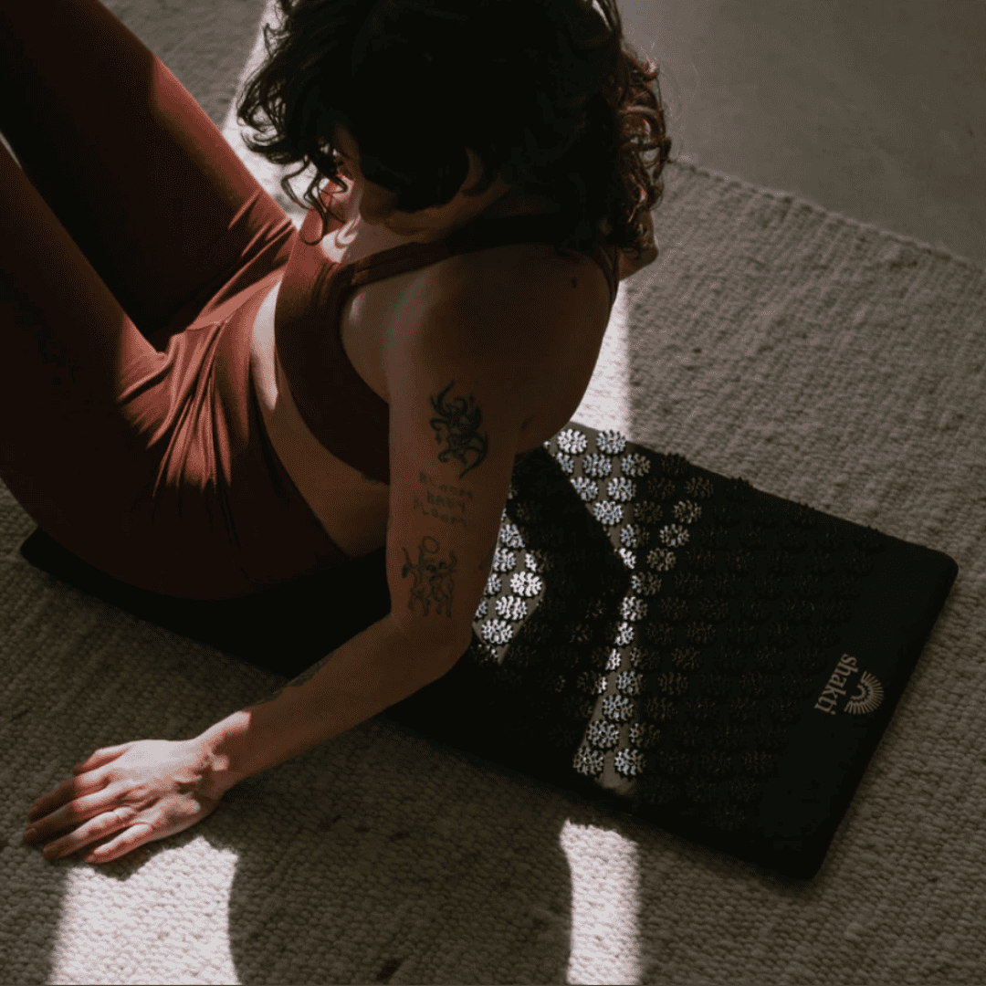 Person sitting on a black acupressure mat with flower-shaped spikes in warm natural sunlight.