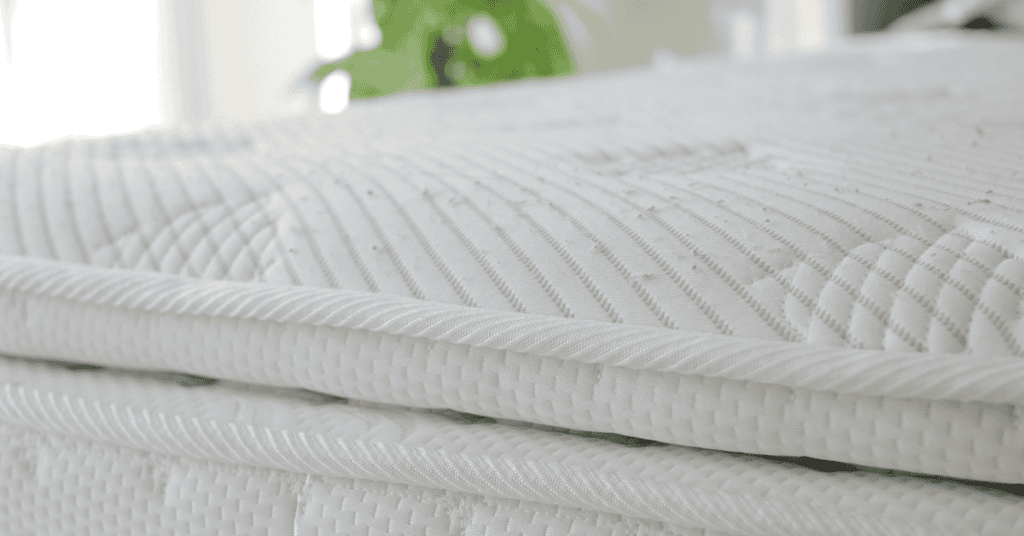 Close-up of a white quilted mattress surface and side seam in a bright bedroom.