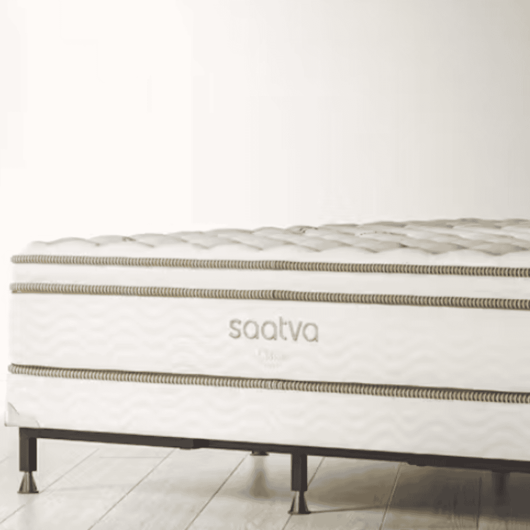Saatva mattress displayed on a simple bed base in a bright, neutral bedroom setting.