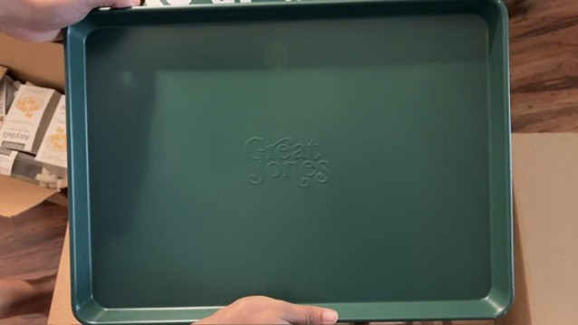 Hands holding a green Great Jones Holy Sheet baking pan with its embossed logo visible