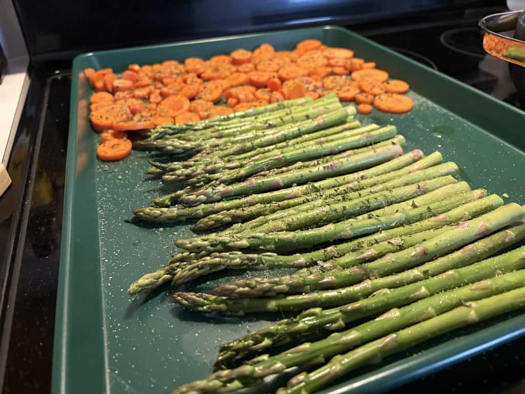 Seasoned asparagus and sliced carrots arranged on a green Great Jones Holy Sheet baking pan