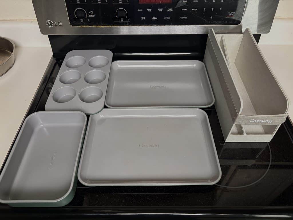 Caraway mini bakeware arranged on a stovetop: two baking sheets, a rectangle pan, a six-cup muffin pan, and an organizer.