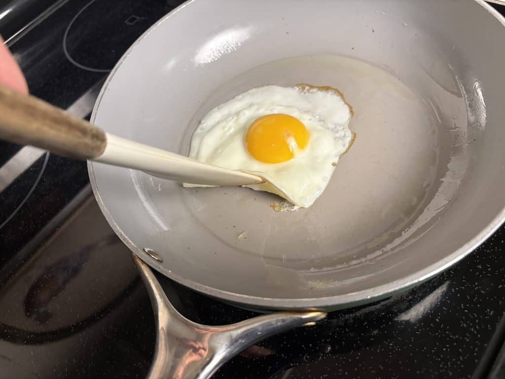 Silicone spatula lifting the edge of a fried egg in a Caraway ceramic pan, with olive oil visible on the cooking surface.
