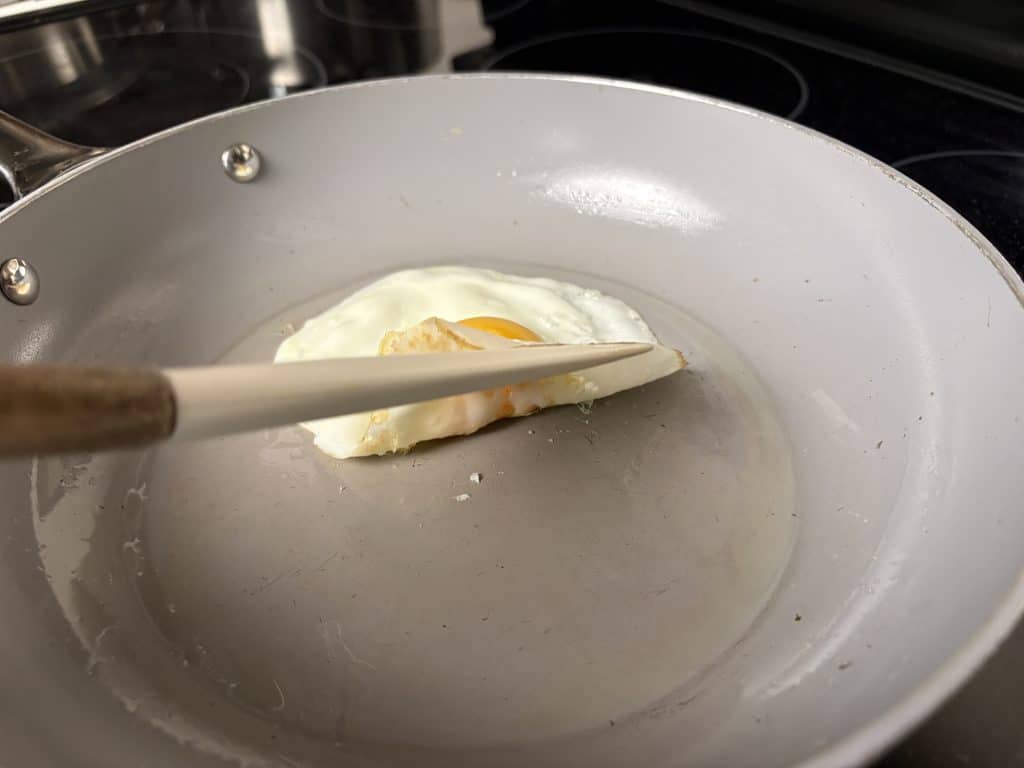 Silicone spatula lifting a fried egg in a Caraway pan, exposing the cooking surface with a few small egg crumbs remaining