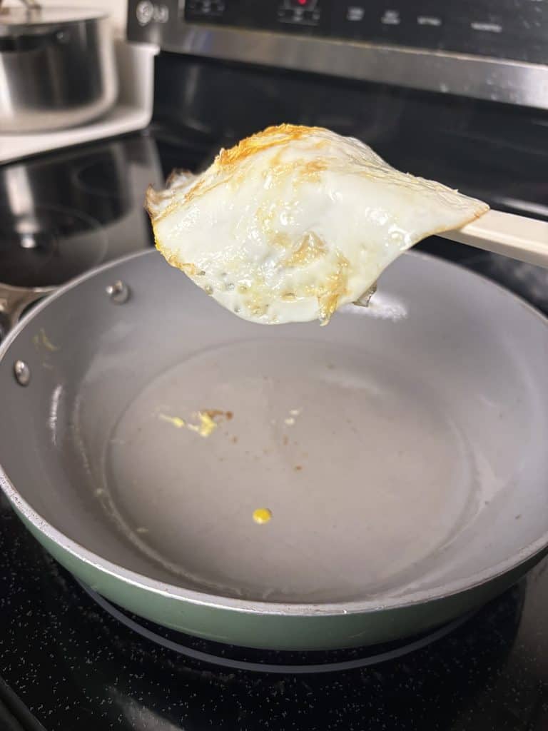 Fried egg held on a silicone spatula above a sage Caraway pan, with a little oil and scattered egg residue inside