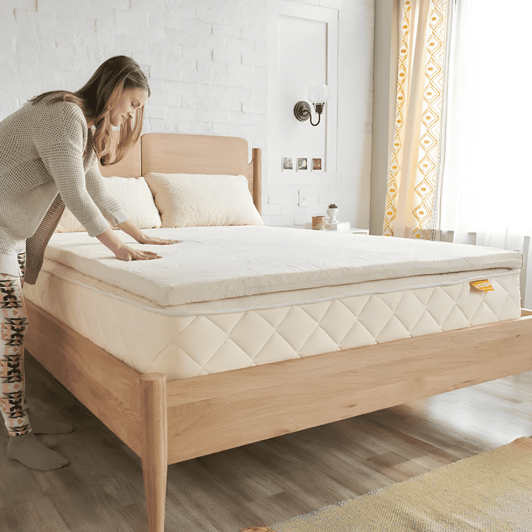 Woman pressing her hands into a white organic mattress topper on a wooden bed frame in a bright bedroom with patterned curtains.