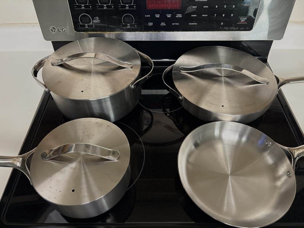 Caraway stainless steel cookware arranged on the stovetop, with lids on the saucepan, larger pot, and sauté pan