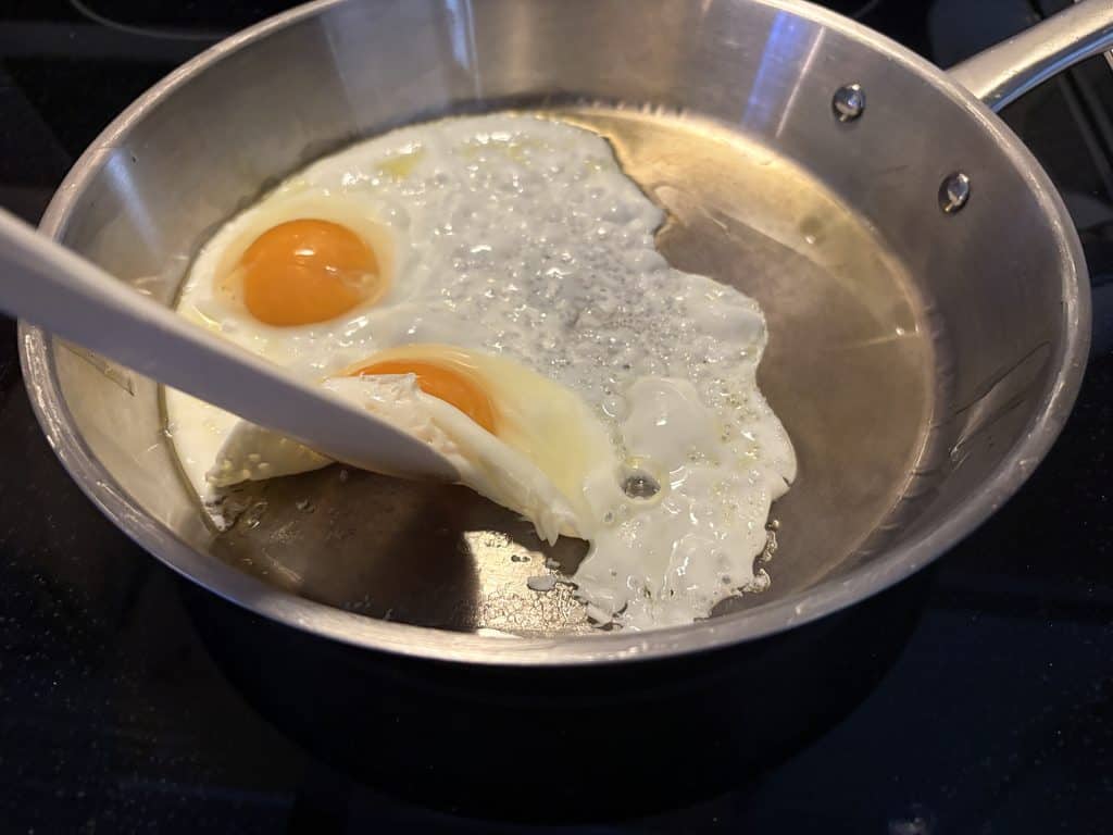 Spatula lifting the edge of one of two fried eggs in a La Sera stainless steel pan, with egg residue underneath