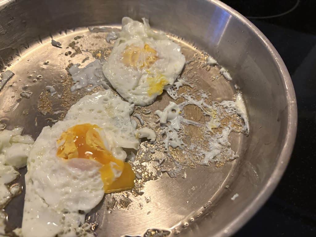 Two torn fried eggs with broken yolks and scattered stuck-on egg white in a La Sera stainless steel pan