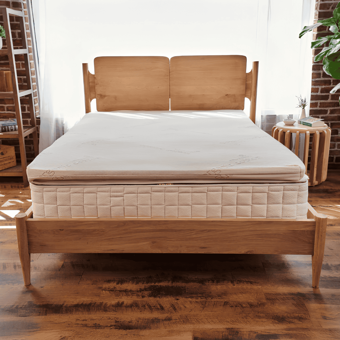 Cream-colored organic latex mattress topper on a quilted mattress with a wooden bed frame in a bright bedroom with exposed brick walls.