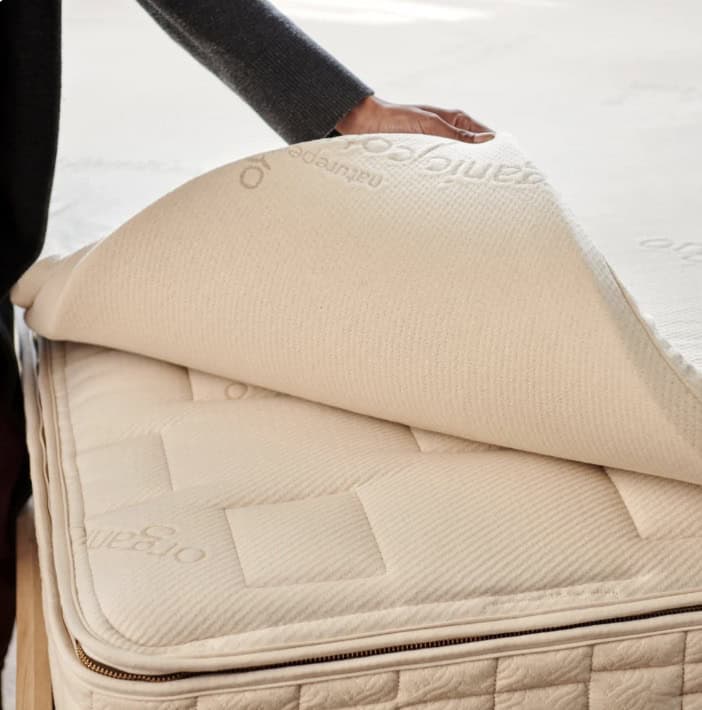 Person lifting the corner of a cream-colored mattress topper to show its thickness and the quilted mattress underneath