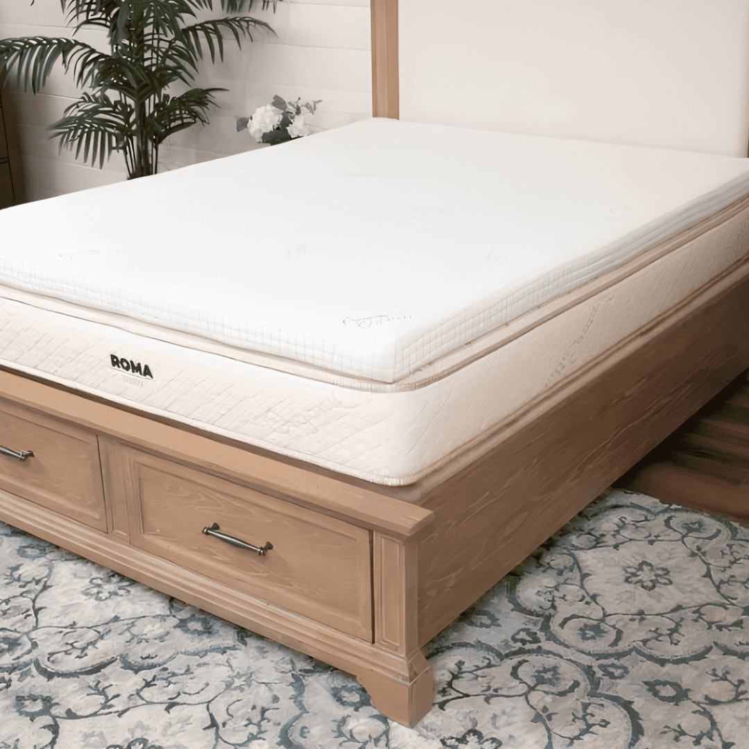 White mattress topper placed on a mattress with a light wooden storage bed frame in a naturally lit bedroom.